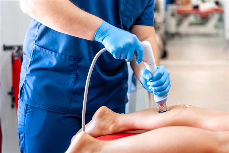 Chiropractor using a handheld shockwave therapy device on a patient's lower leg.
