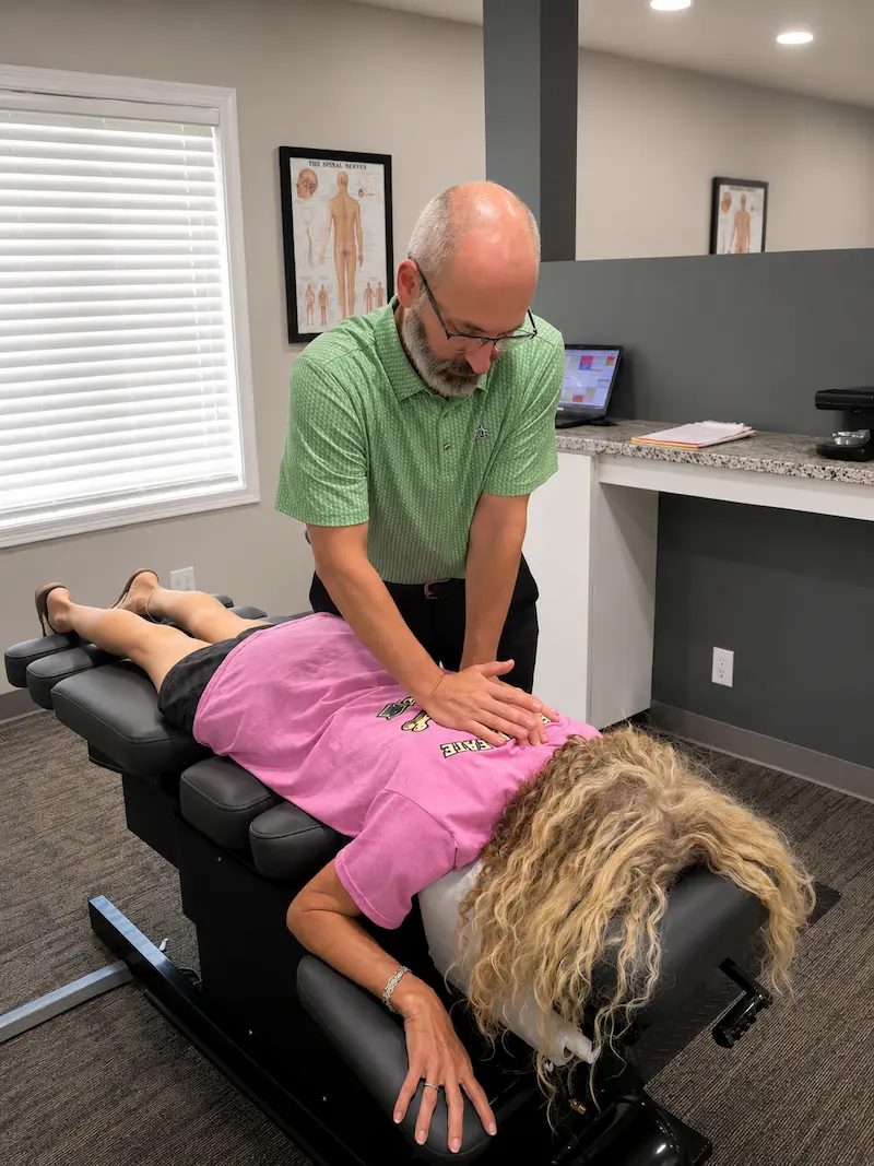 A male chiropractor adjusts a female patient's upper back on a black chiropractic table in a clinic.